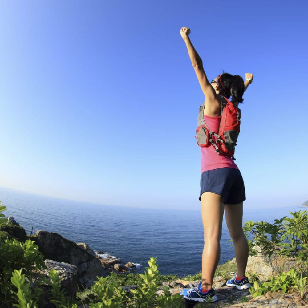 cheering young fitness woman open arms on seaside mountain peak